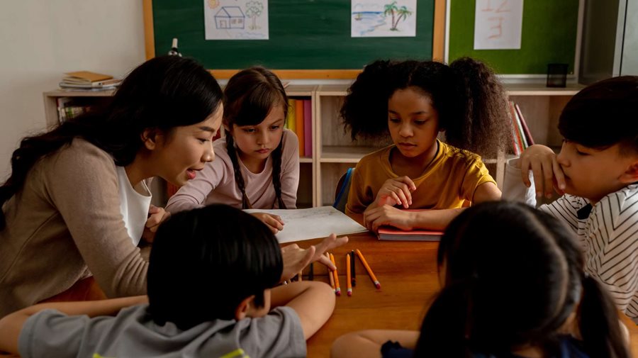 Female teacher of Vietnamese descent sits at a table teaching a group of 5 students of varying ethnicities using colored pencils.