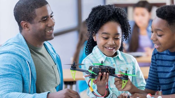 Black educator smiles at two STEM students building a drone in a classroom.